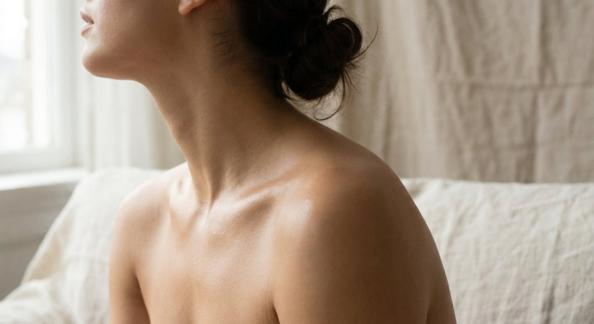 A softly lit close-up of a woman's glowing, calm skin on her shoulder and neck against a warm neutral background.