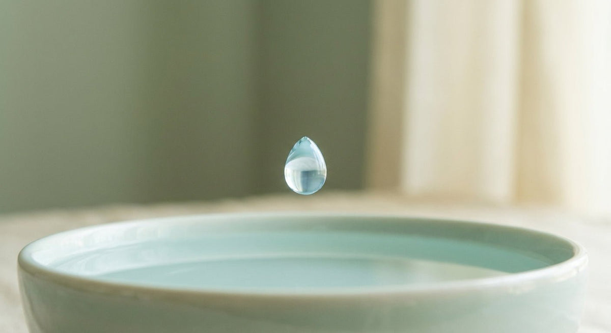A single luminous water droplet suspended above a still pool surface, photographed in soft natural light against a muted sage and ivory background.
