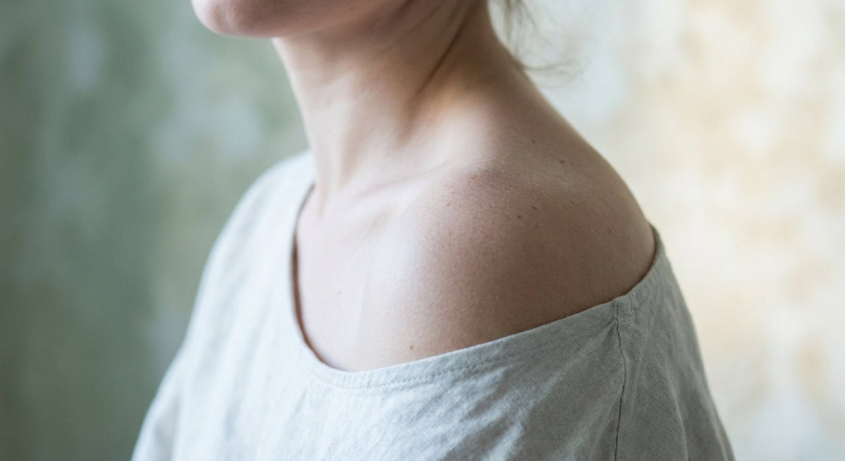 A close-up editorial photograph of bare skin on a woman's shoulder and neck, softly lit in cool natural light against a muted pale background, evoking sensitivity and skin vulnerability.